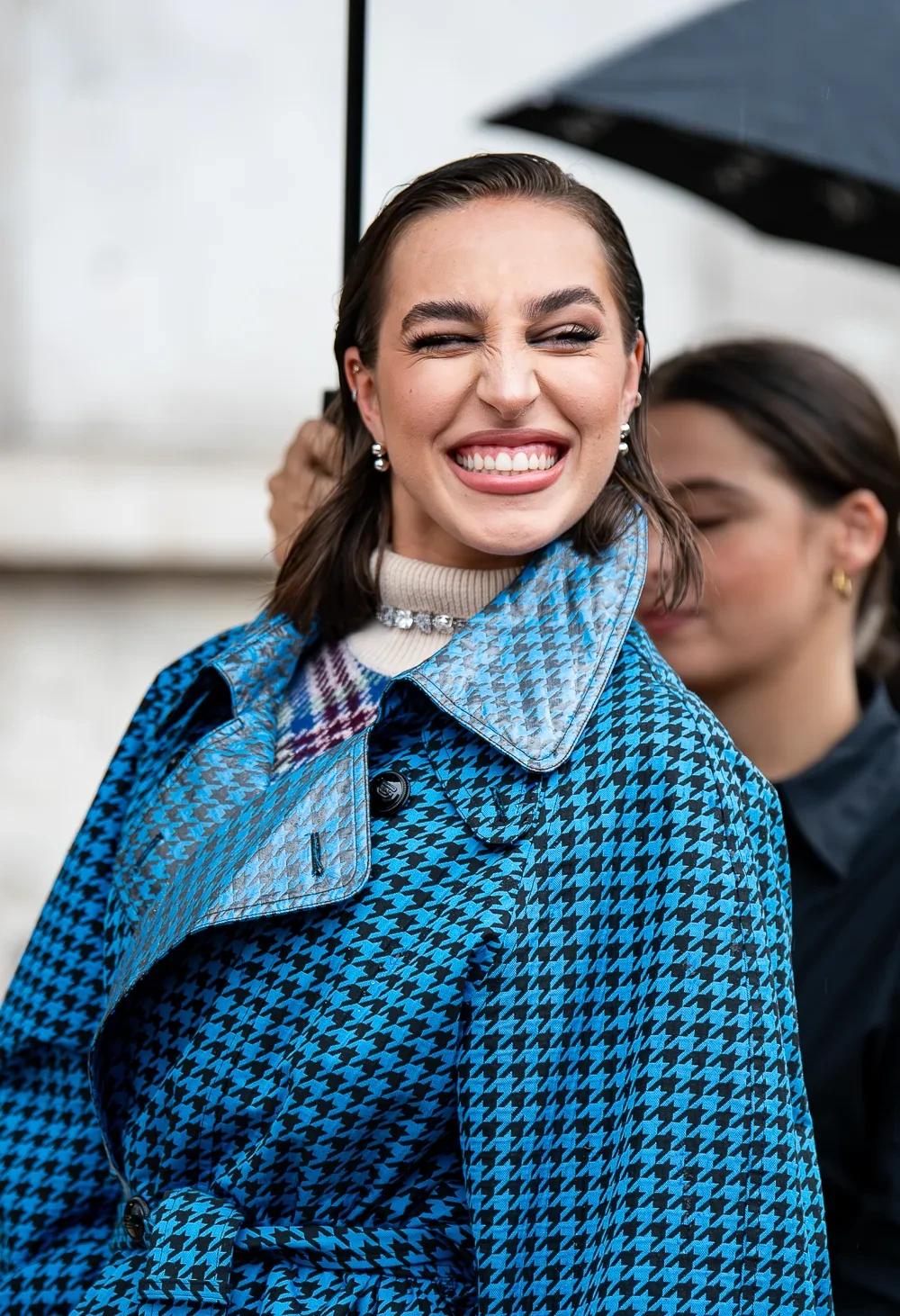 Ginevra Mavilla éclatant de rire, en manteau pied-de-poule bleu à col vernis, sous un parapluie au défilé Rabanne