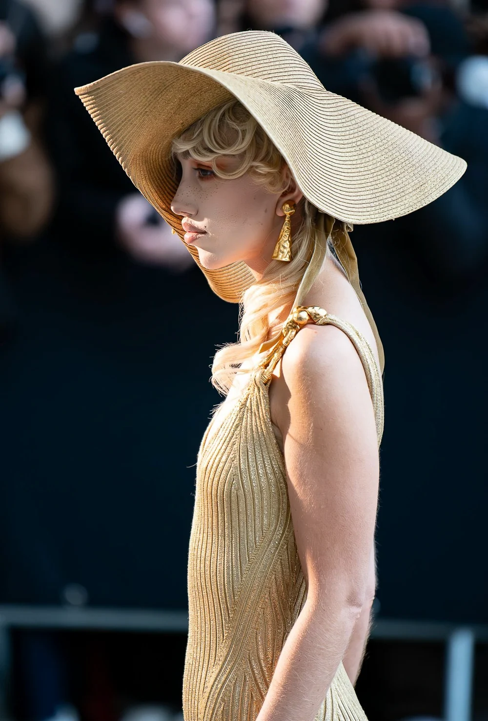 Nadia Lee Cohen de profil sous une capeline de paille, en robe de maille dorée, au défilé Schiaparelli à Nice