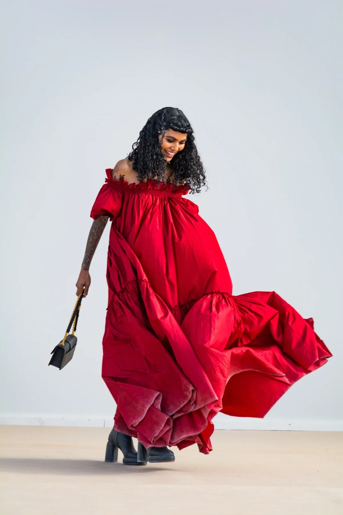 Sophia Roe en ample robe rouge à épaules dénudées qui virevolte, sac noir à la main, au défilé Dior à Monaco