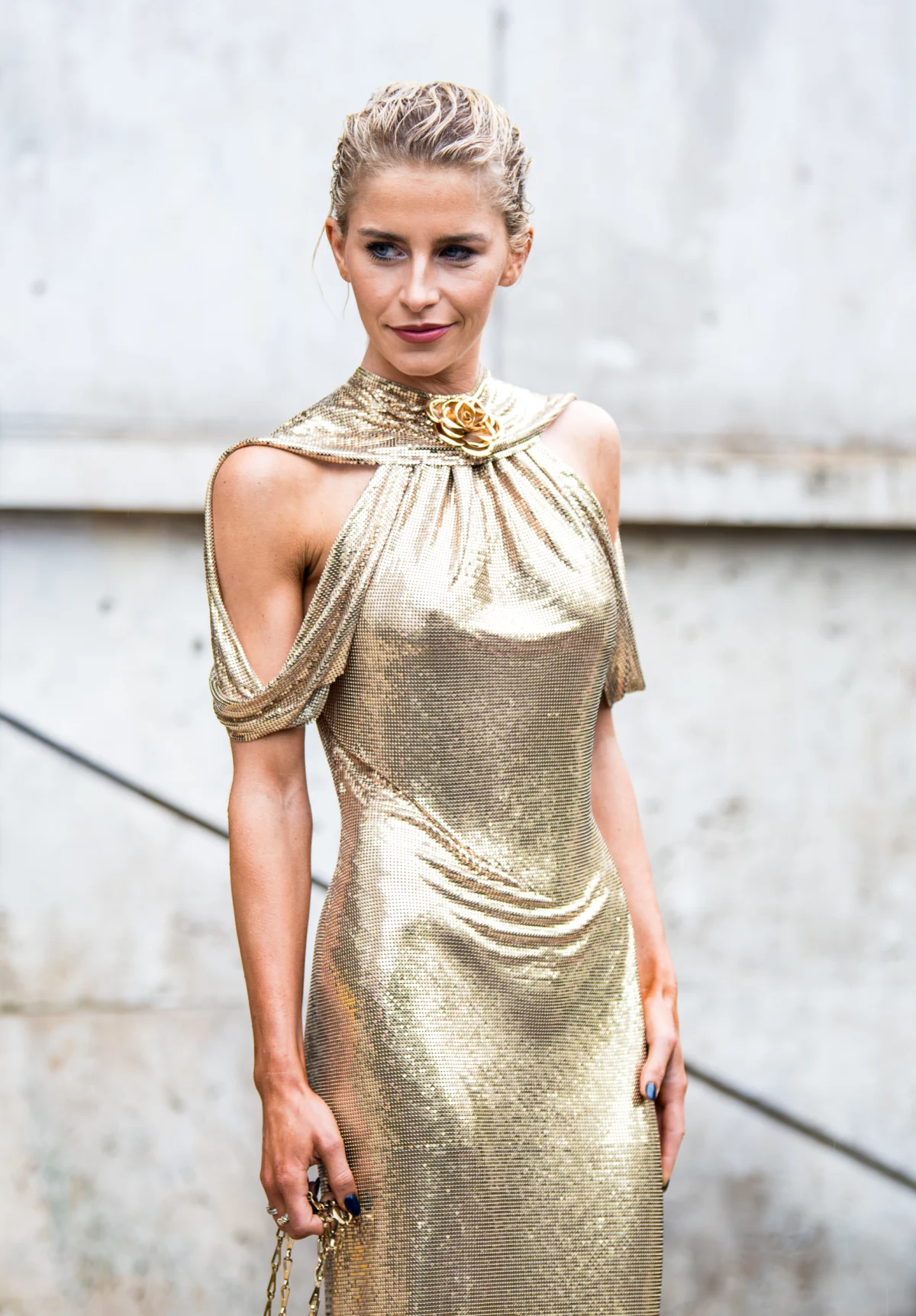 Femme blonde aux cheveux plaqués en robe drapée en lamé doré, broche fleur au col, devant un mur de béton à Cannes