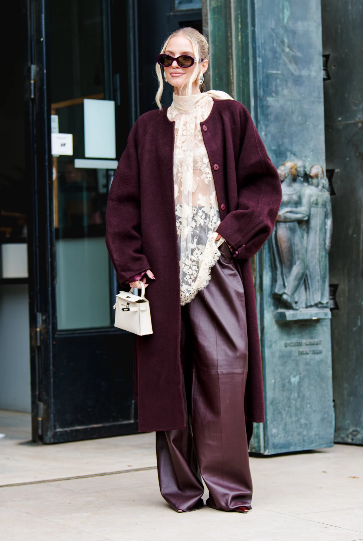 Femme blonde en lunettes noires, long manteau bordeaux, blouse en dentelle et pantalon en cuir, devant une entrée à Cannes