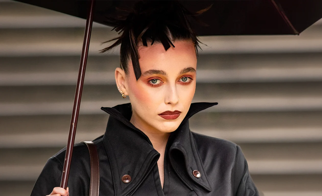 Femme aux cheveux noirs coiffés en pics sous un parapluie, trench en cuir noir col relevé, rouge à lèvres bordeaux
