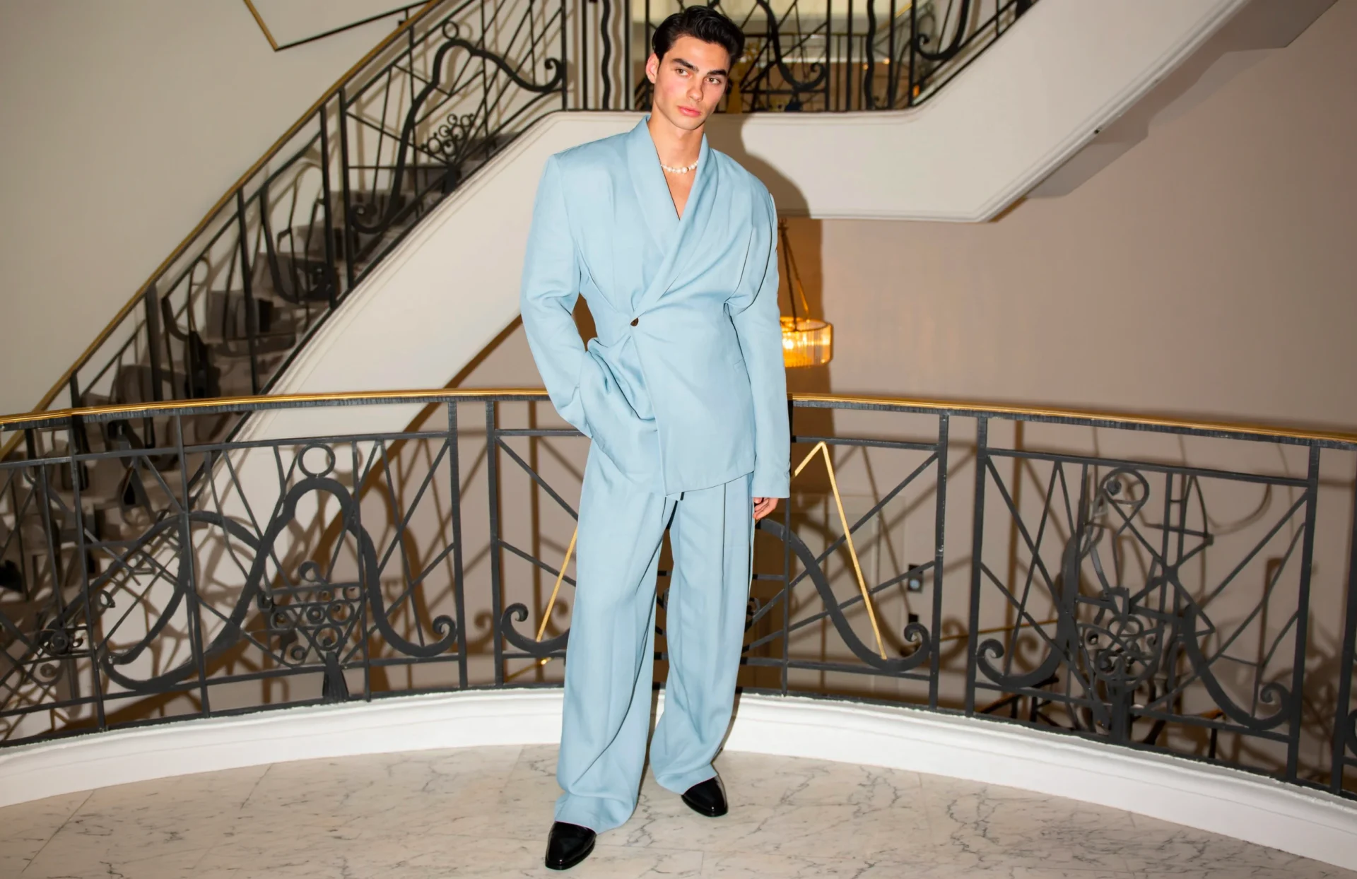 Homme en costume bleu ciel posant dans un escalier d'hôtel de prestige en marbre et ferronnerie dorée