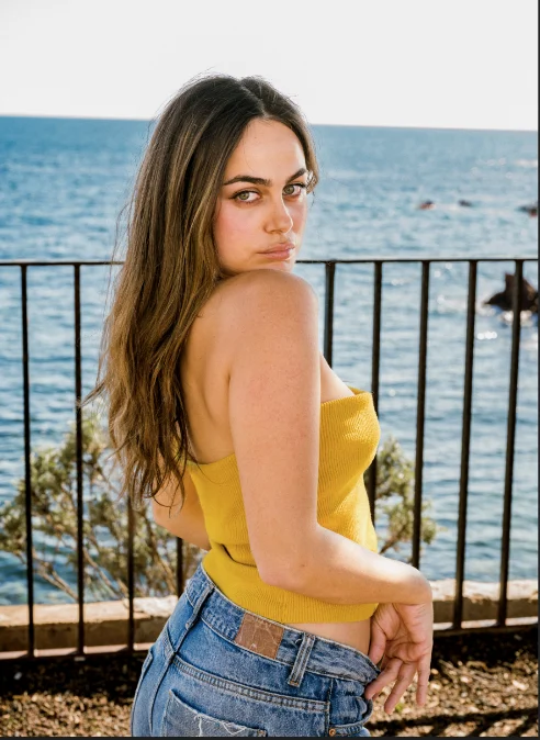 Jeune femme en bustier jaune et jean, regard par-dessus l'épaule, contre une rambarde face à la mer, à Cannes