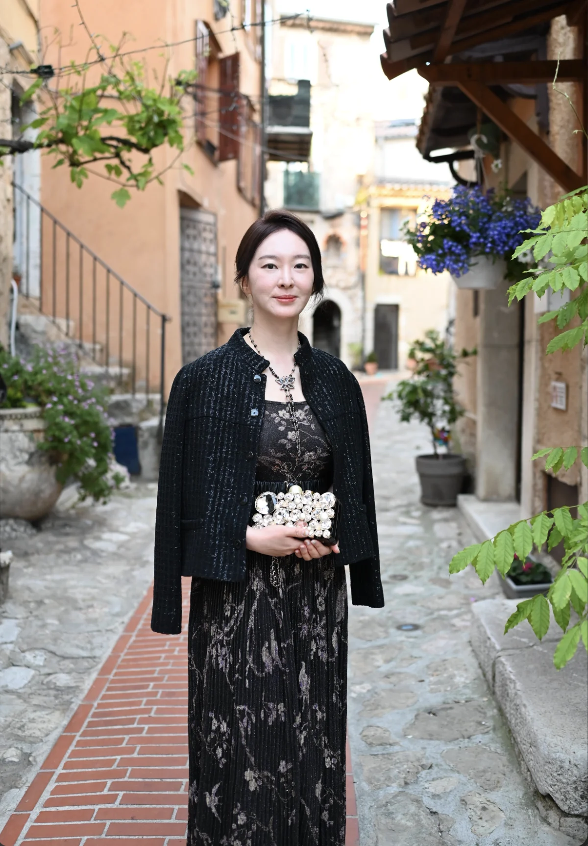 Femme en veste en tweed noire et robe longue imprimée, pochette perlée à la main, dans une ruelle fleurie d'Èze
