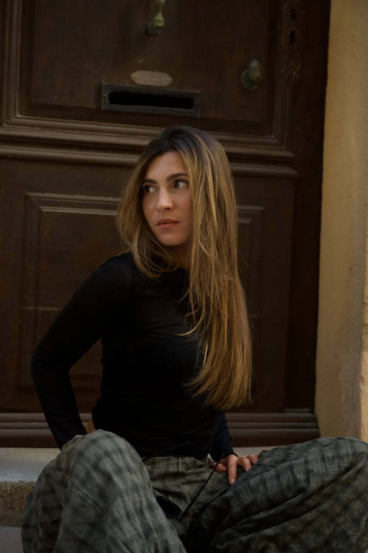 Jeune femme aux longs cheveux méchés en pull noir, regard de côté, assise devant une porte en bois à Saint-Tropez