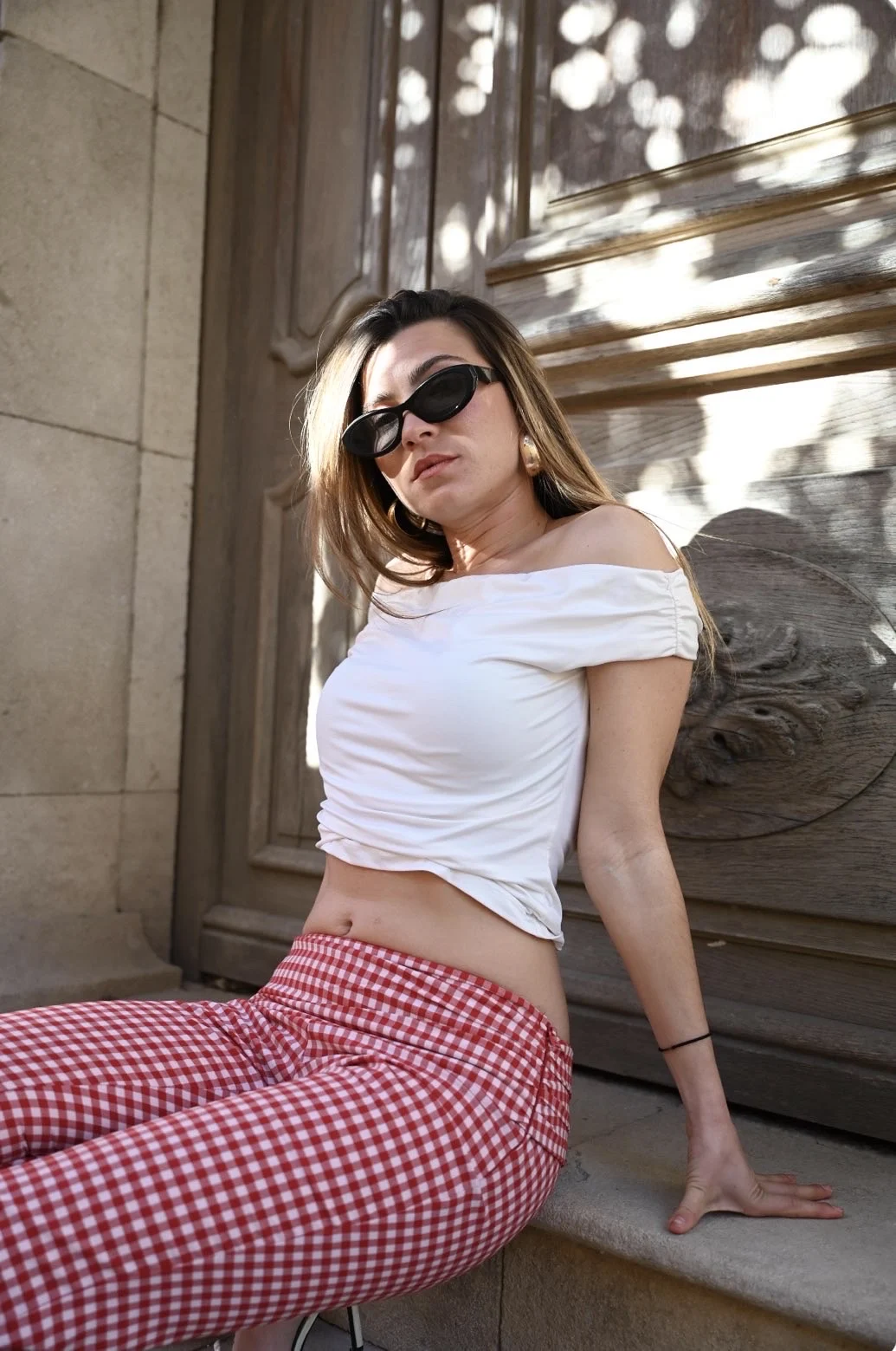 Jeune femme en haut blanc, lunettes de soleil et pantalon vichy rouge, assise contre une porte sculptée à Saint-Tropez
