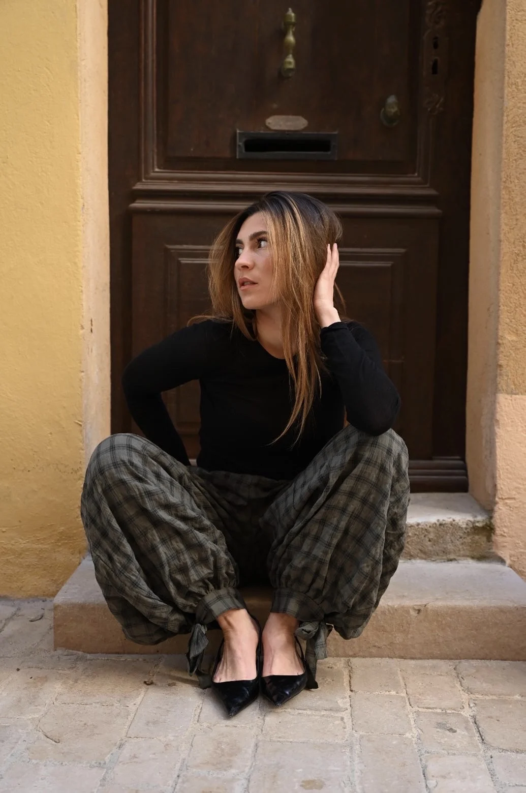 Jeune femme en pantalon à carreaux, assise sur le seuil d'une porte en bois, la main dans les cheveux, à Saint-Tropez