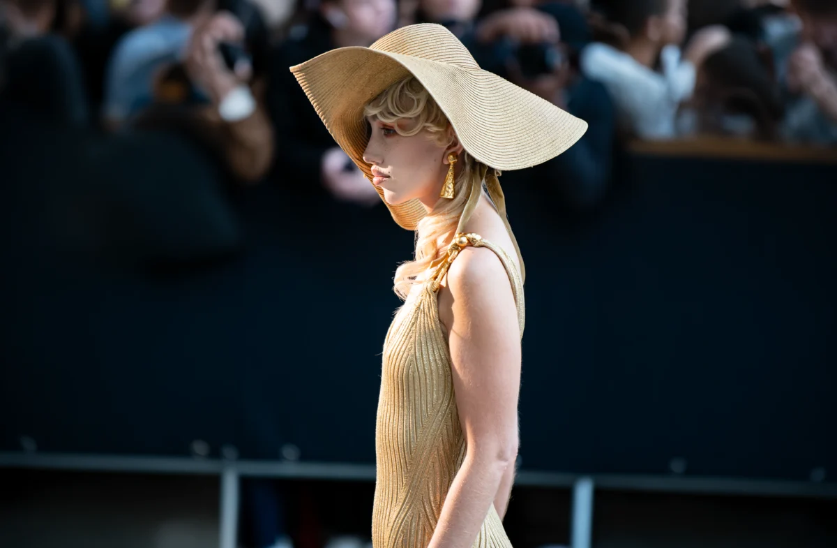 Femme de profil sous une grande capeline de paille, robe en maille côtelée dorée et boucles en or, devant les photographes