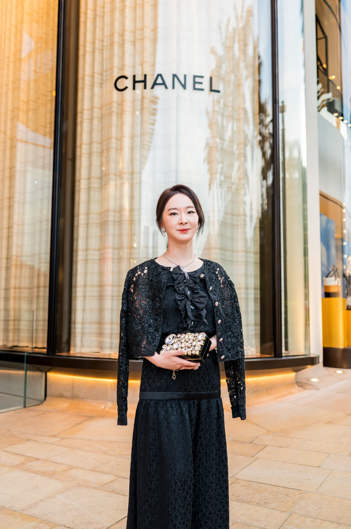 Femme en veste et jupe longue en dentelle noire, pochette bijou à la main, devant la boutique Chanel à Monaco