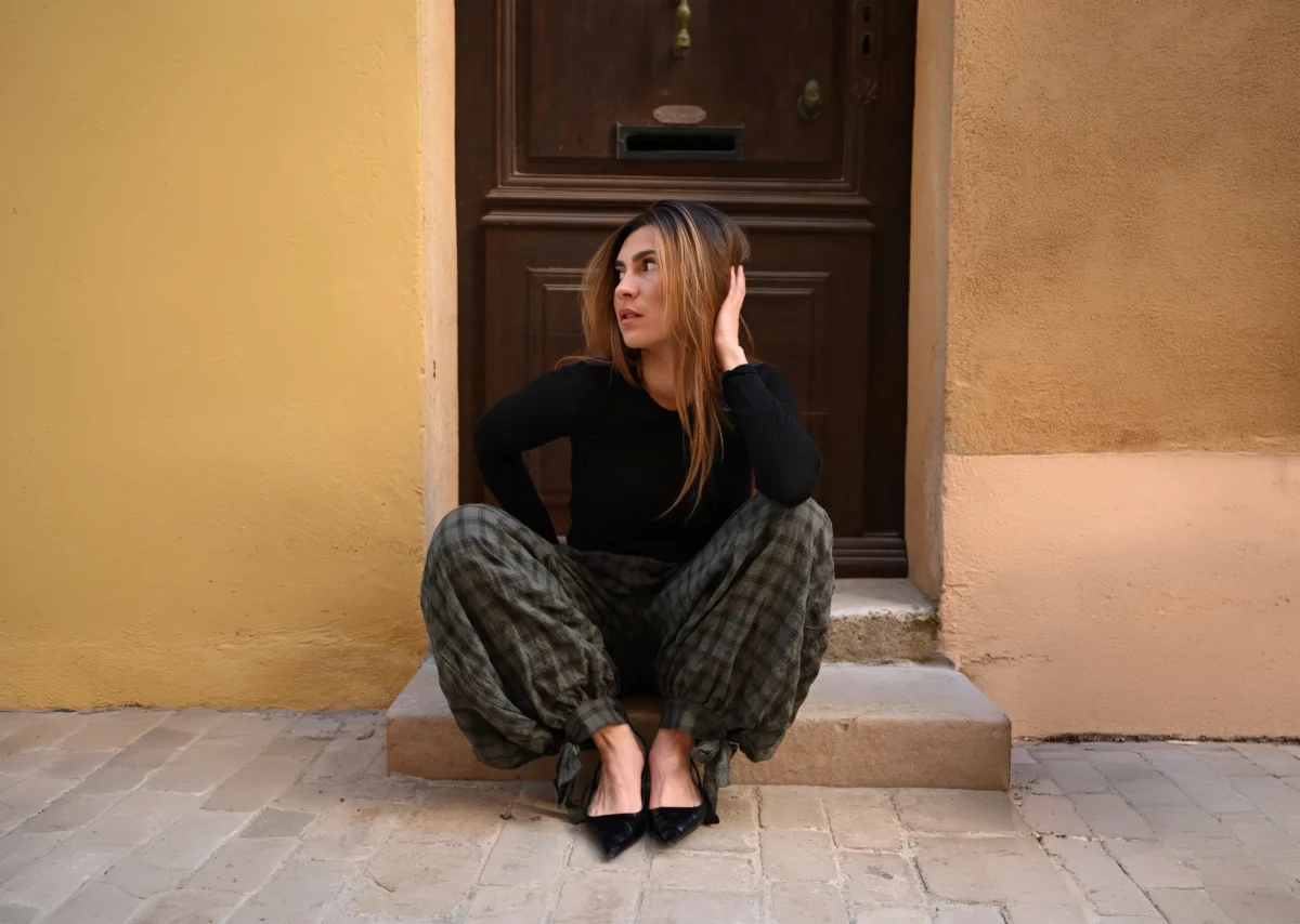 Jeune femme en haut noir et pantalon à carreaux, assise sur le seuil d'une porte ancienne en bois, la main dans les cheveux